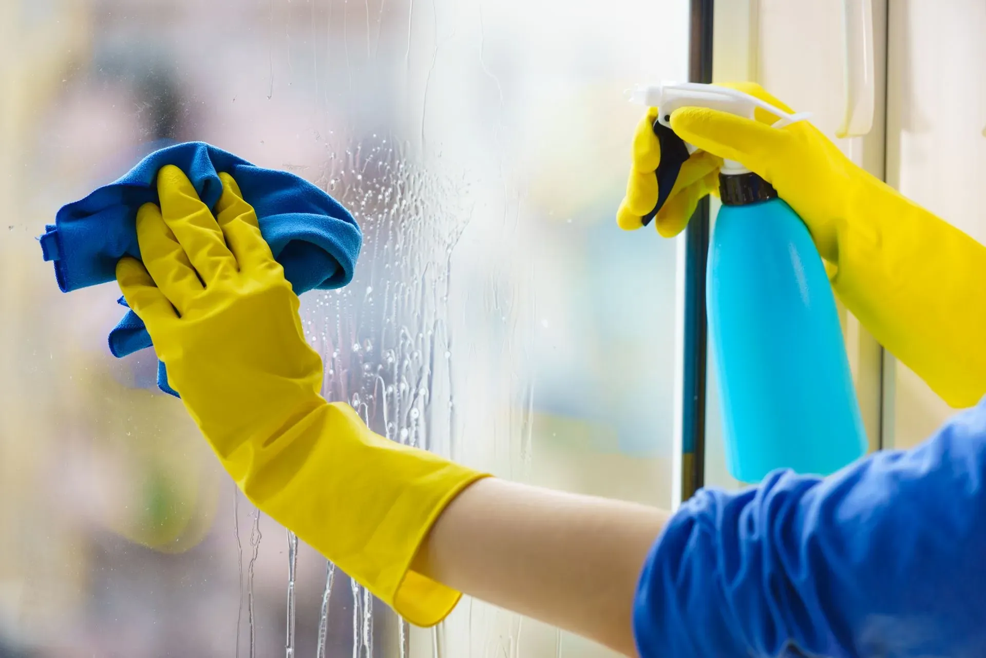 A person wearing yellow gloves is cleaning a window with a cloth and spray bottle.