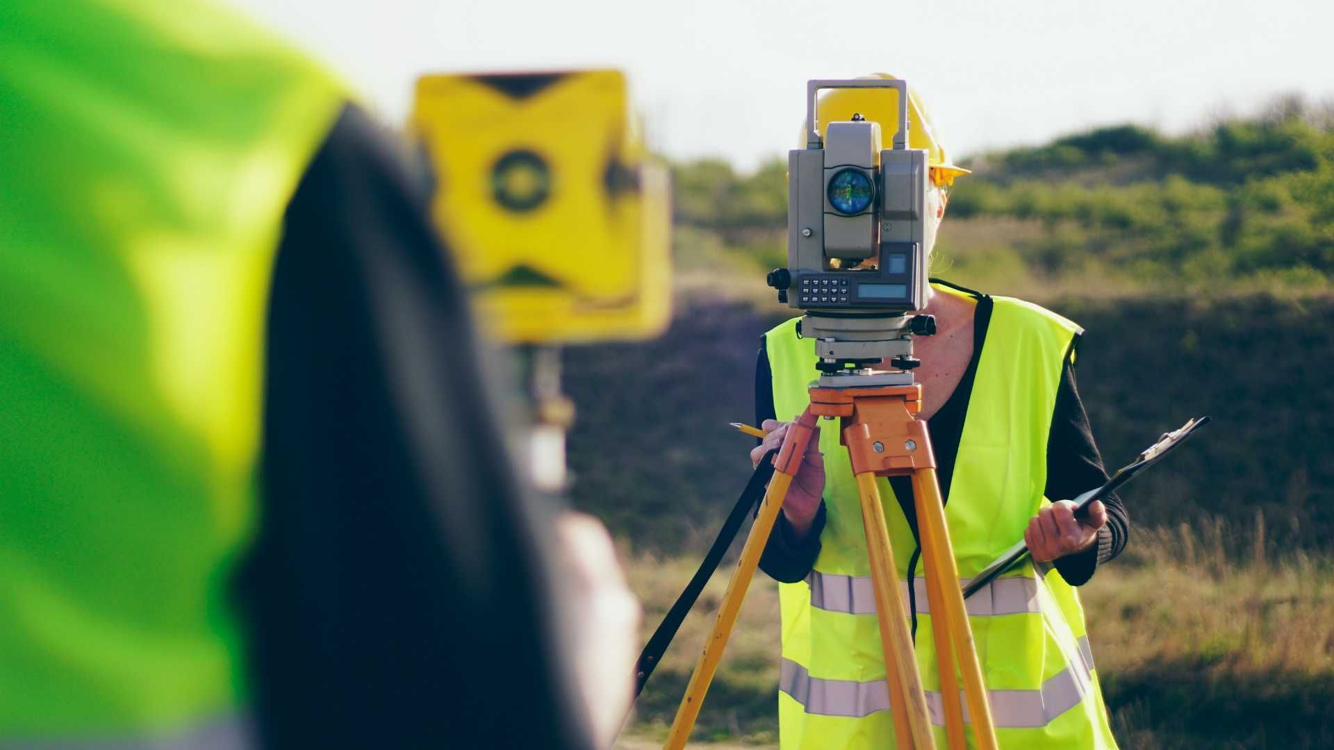 A man in a yellow vest is using a theodolite on a tripod.