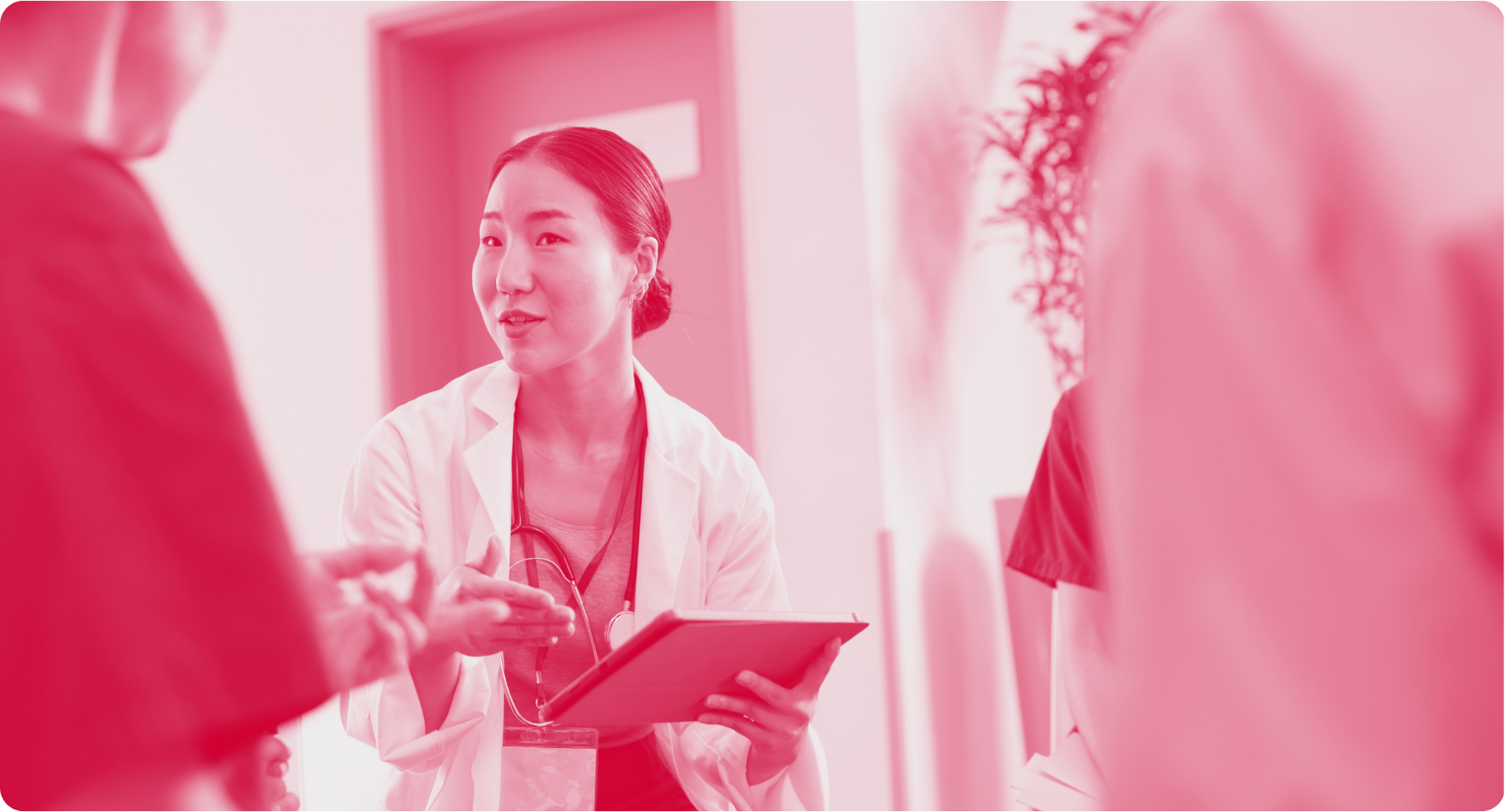 Doctor in a white coat holding a tablet, speaking with people in a hallway.