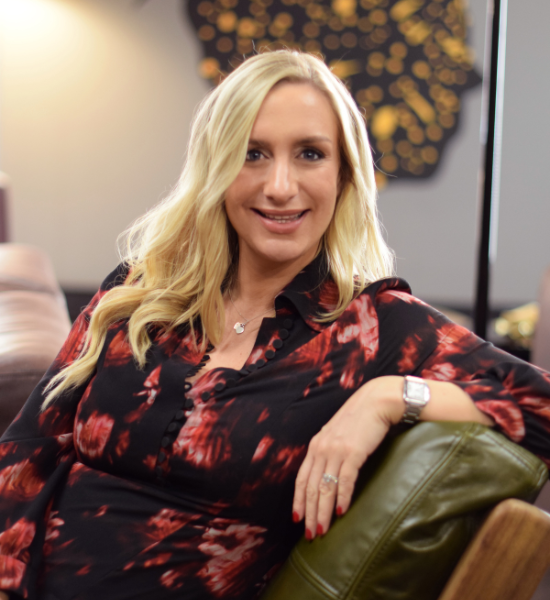 Woman with blonde hair, smiling, wearing patterned top, resting arm on green chair, indoors.
