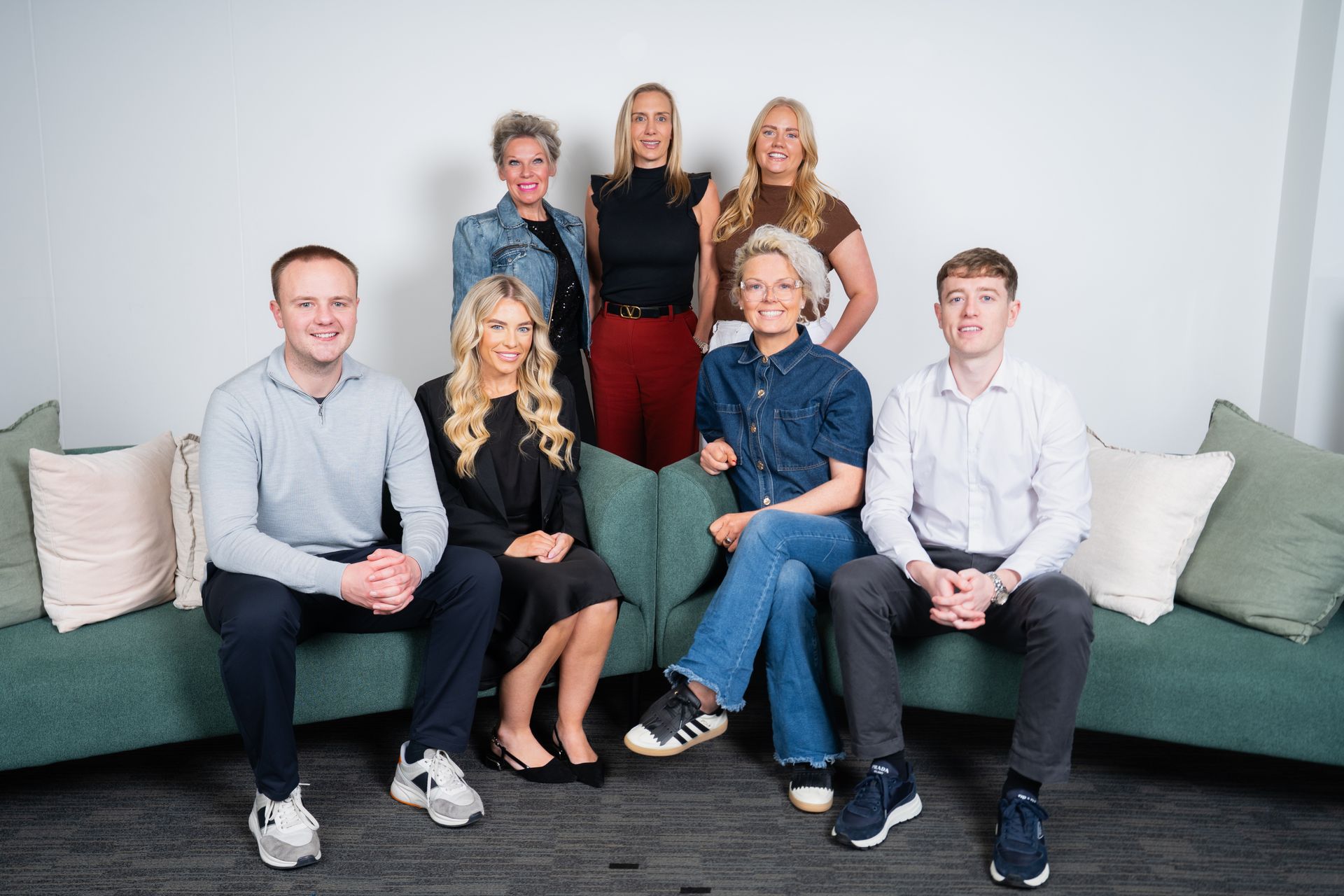 Group of nine people posing on and around a green couch against a white wall; some are smiling.