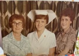 Three women pose for a photo: One in a nursing uniform, the others in patterned clothing, set against a floral curtain.