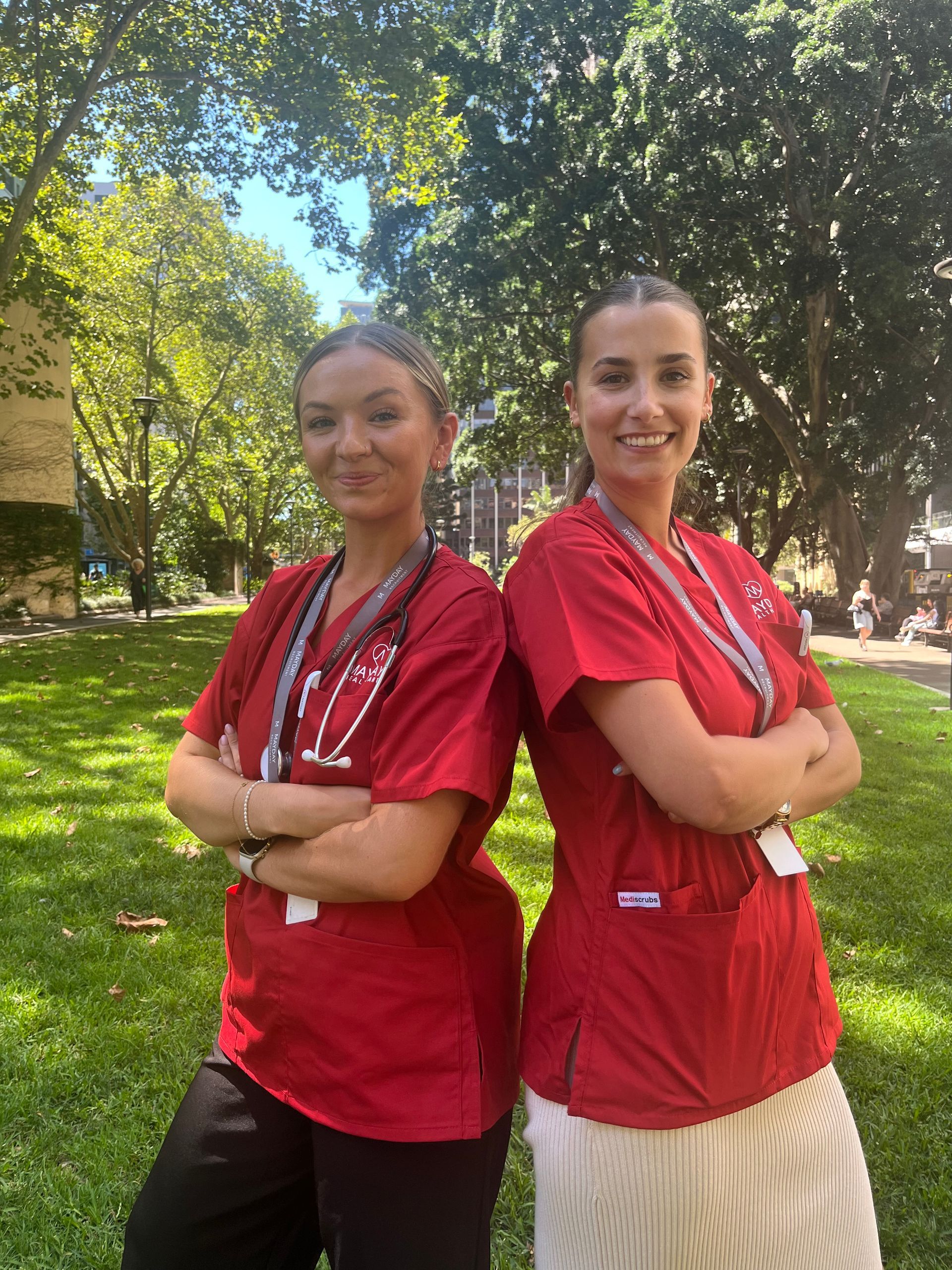 Two women in red scrubs stand back-to-back, arms crossed, in a green outdoor setting.