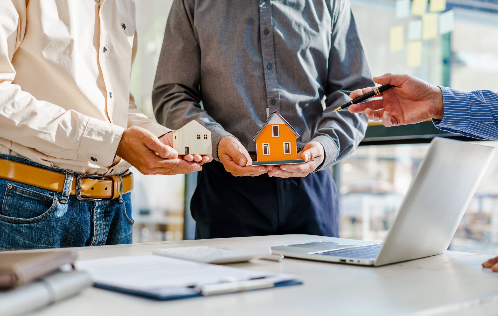Three people in an office hold house models and keys across a desk with a laptop.