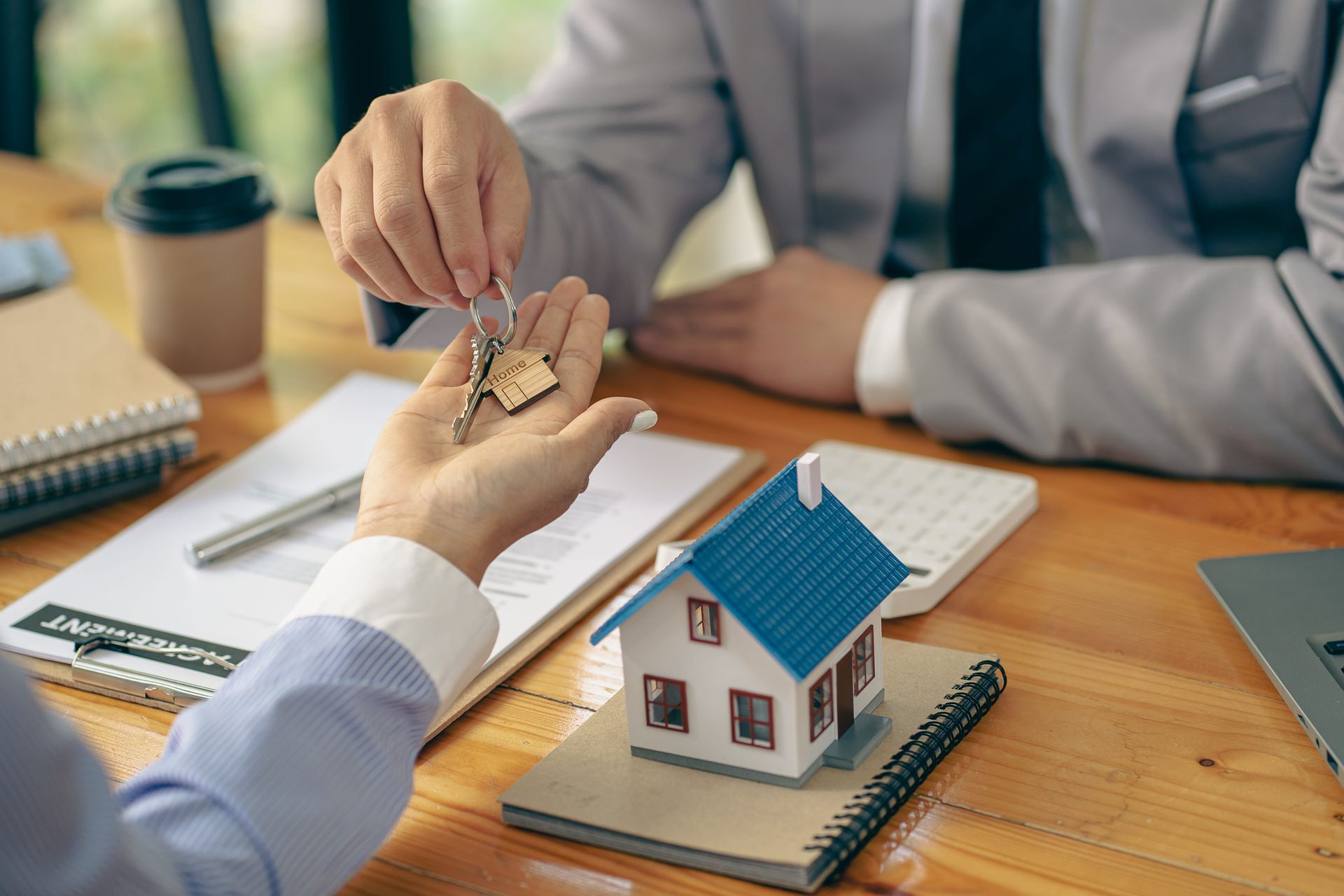 Hands exchanging house keys over a desk with a small model house and paperwork