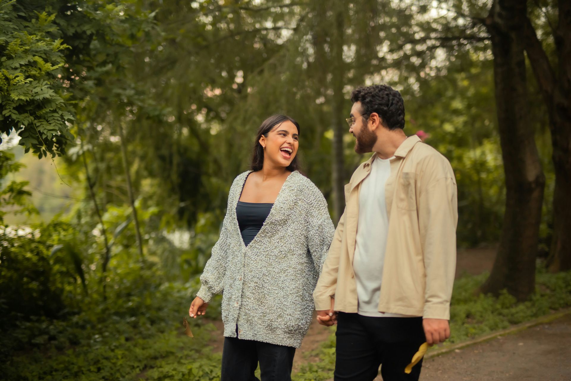 A smiling couple holds hands, walking on a path in a park. Green trees surround them.
