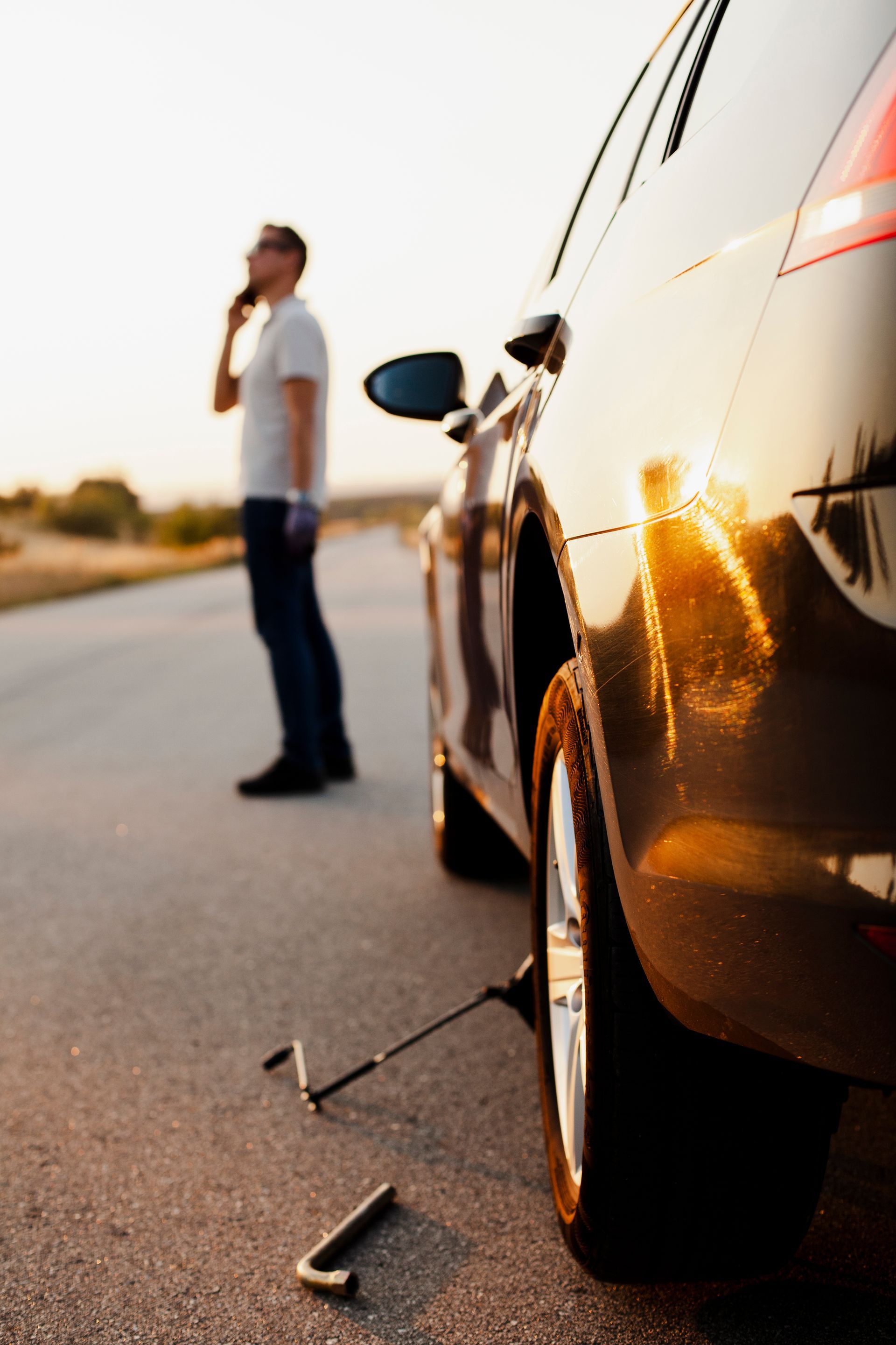 Broken car on roadside with a man standing nearby, speaking on a phone at sunset