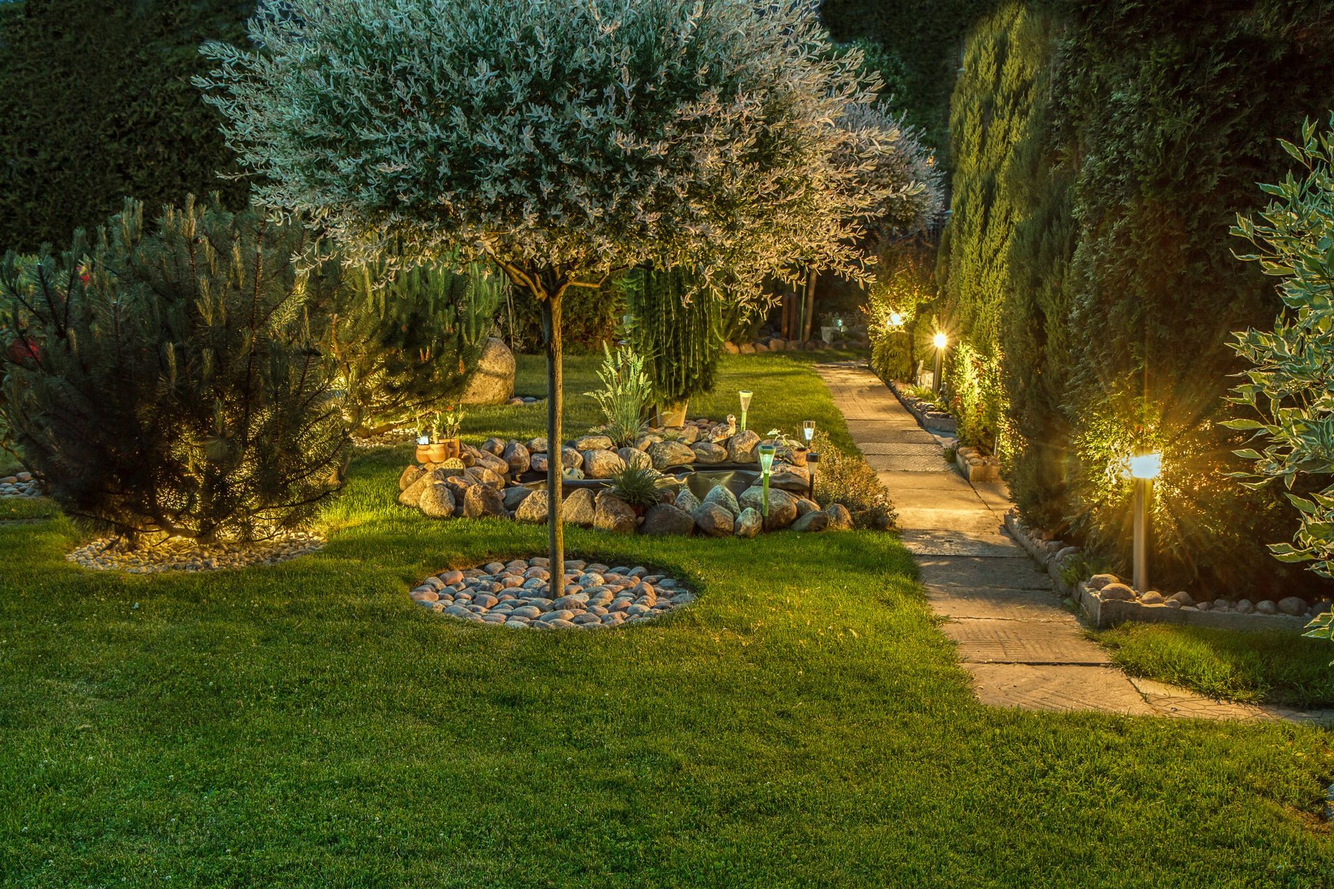 Nighttime garden with lit pathways, trimmed lawn, and trees surrounding a small seating area Nighttime garden with lit pathways, trimmed lawn, and trees surrounding a small seating area