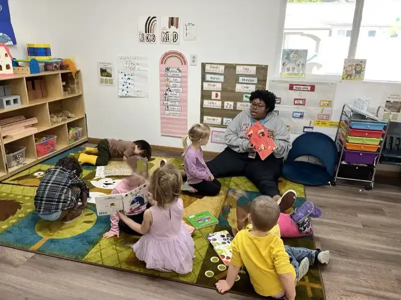 An adult sitting on a colorful rug reads a book to a group of children in a brightly decorated classroom.