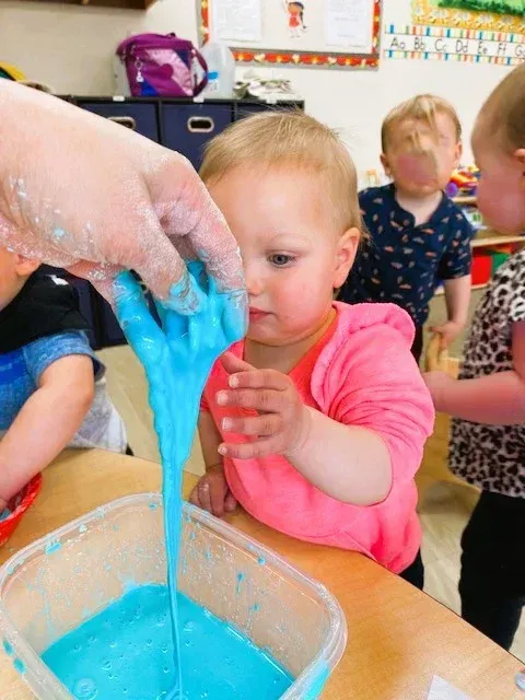 A toddler in a pink shirt touches a string of blue slime held by an adult over a plastic container in a classroom.