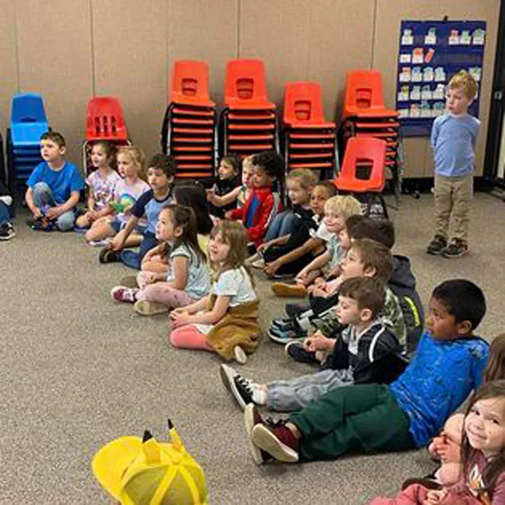 A group of children sit on a carpeted floor in a classroom, listening during an indoor activity near stacked chairs.