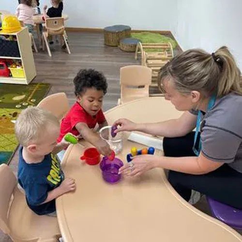 A teacher and two children sit at a table, sorting colorful plastic pieces into small containers during an activity.