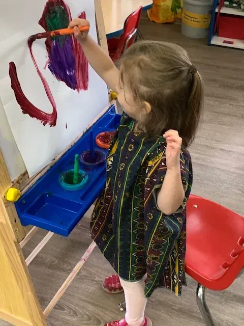 A child wearing a patterned apron paints a large red stroke on a white paper attached to an easel in a classroom.