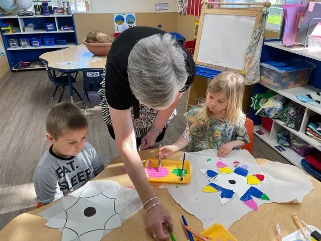 An adult and two children at a table painting star-shaped paper cutouts in a classroom setting.