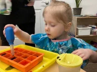 A child using a blue dropper to transfer liquid into an orange silicone ice cube tray on a yellow tray.