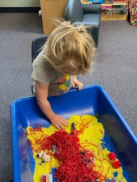 A toddler plays in a sensory bin filled with yellow rice and red yarn, featuring small toy figures in a classroom.