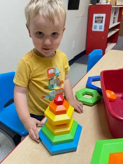 A young child in a yellow t-shirt builds a colorful hexagonal stacking toy on a table in a classroom setting.