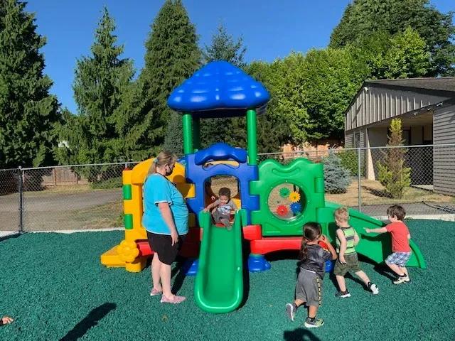 A teacher supervises children playing on a colorful plastic playground structure on a green rubber-surfaced area outdoors.