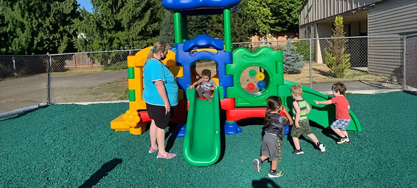 A person supervises children playing on a colorful plastic playground structure on a green rubber-surfaced play area.