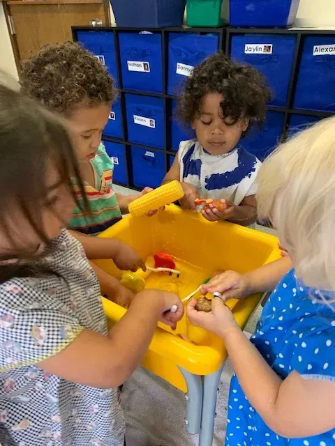 Four children gather around a yellow sensory table in a classroom, engaged in play with various toys.