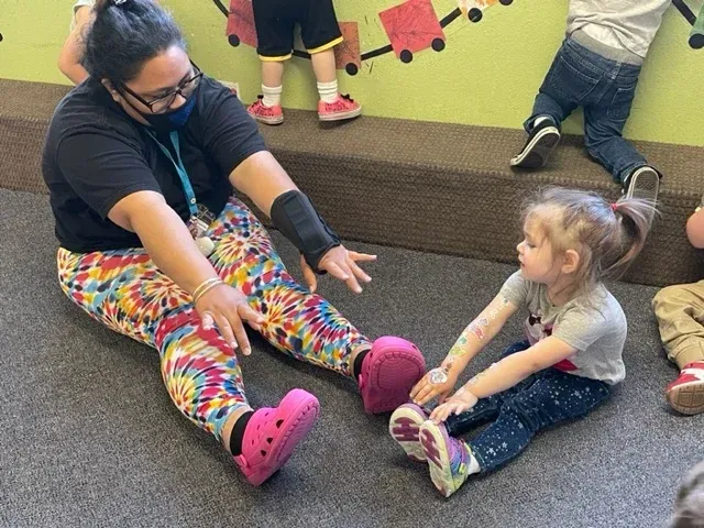 An adult and a child sitting on a carpeted floor, reaching forward toward each other during an activity in a classroom.