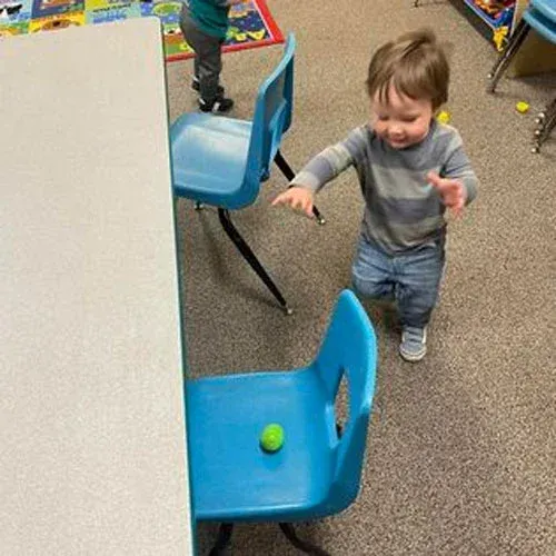 A child in a striped shirt reaches toward a green ball sitting on a blue plastic chair next to a table in a room.