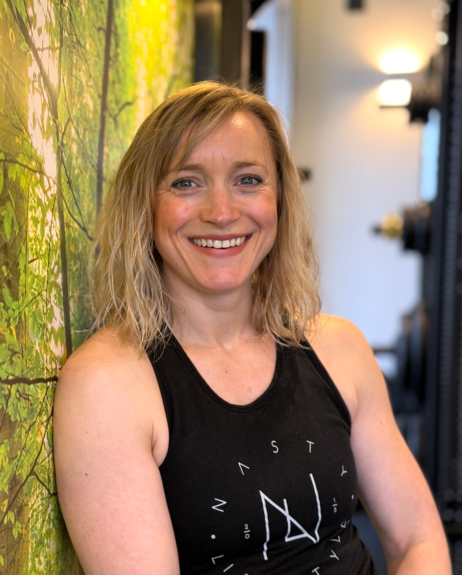 A woman in a black tank top is smiling and leaning against a wall.