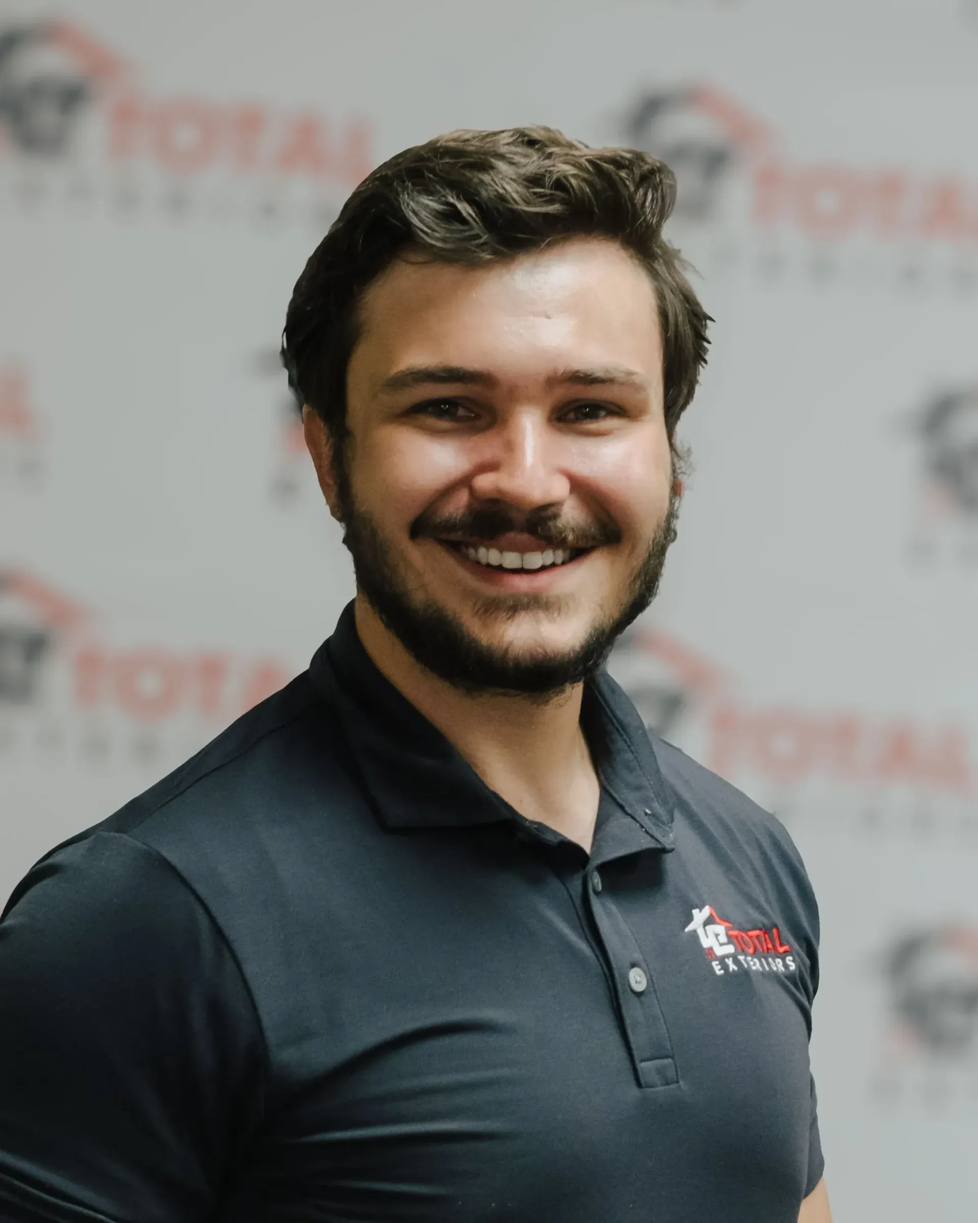 A man with a beard is wearing a black polo shirt and smiling.