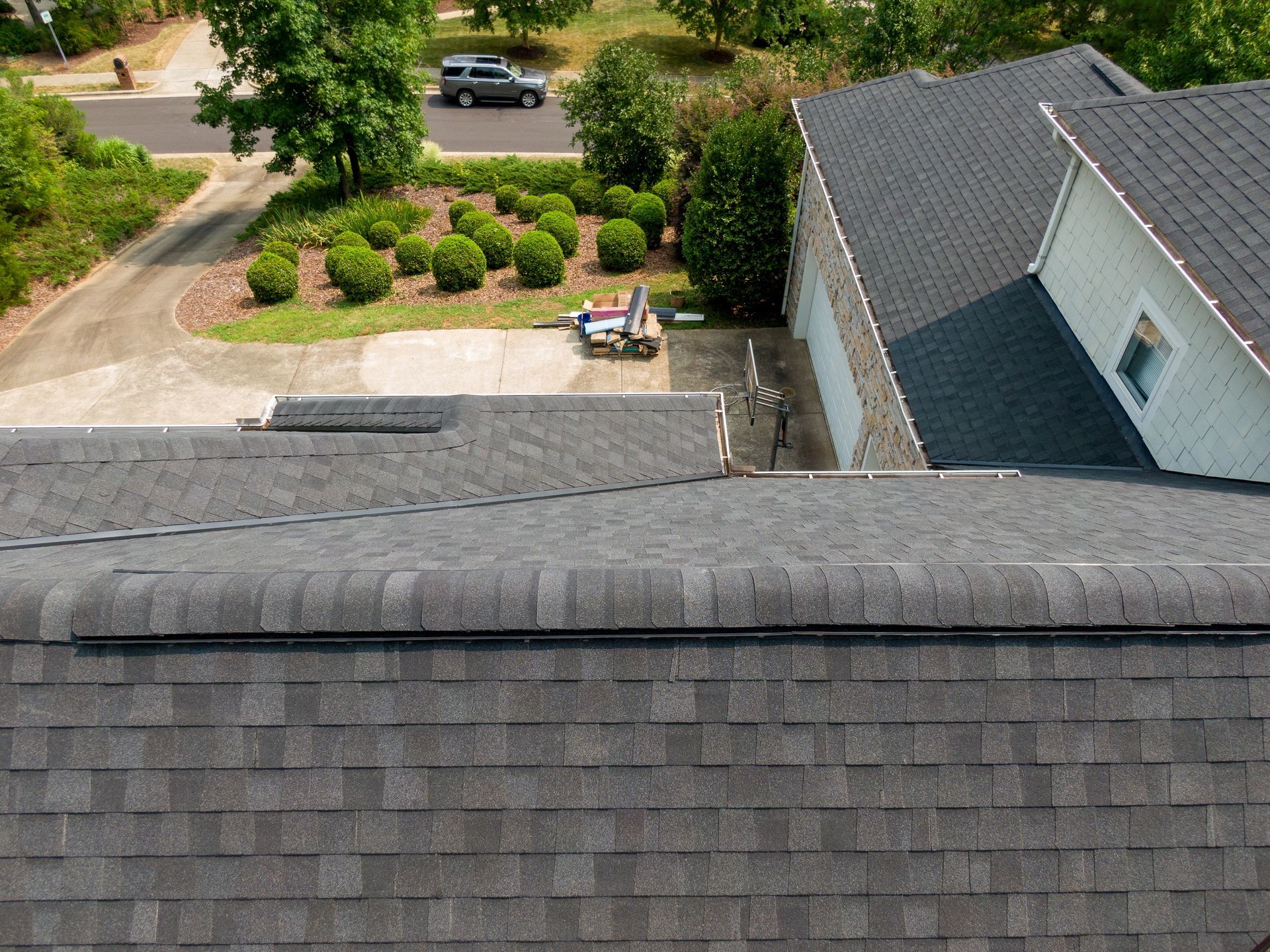 Overhead view of a dark gray shingle roof on a house, with a driveway and landscaped yard visible.