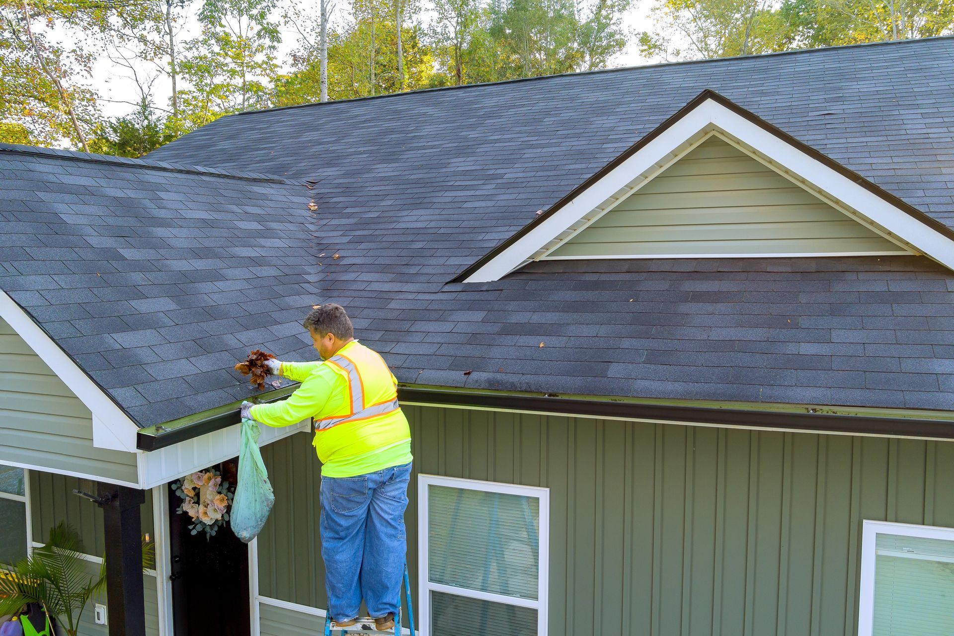 A man is standing on a ladder cleaning the roof of a house.