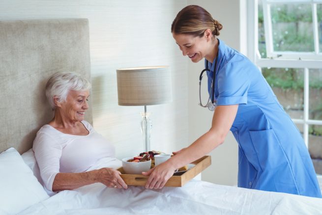 Nurse serving breakfast to a patient in bed; both are smiling in a bright room.