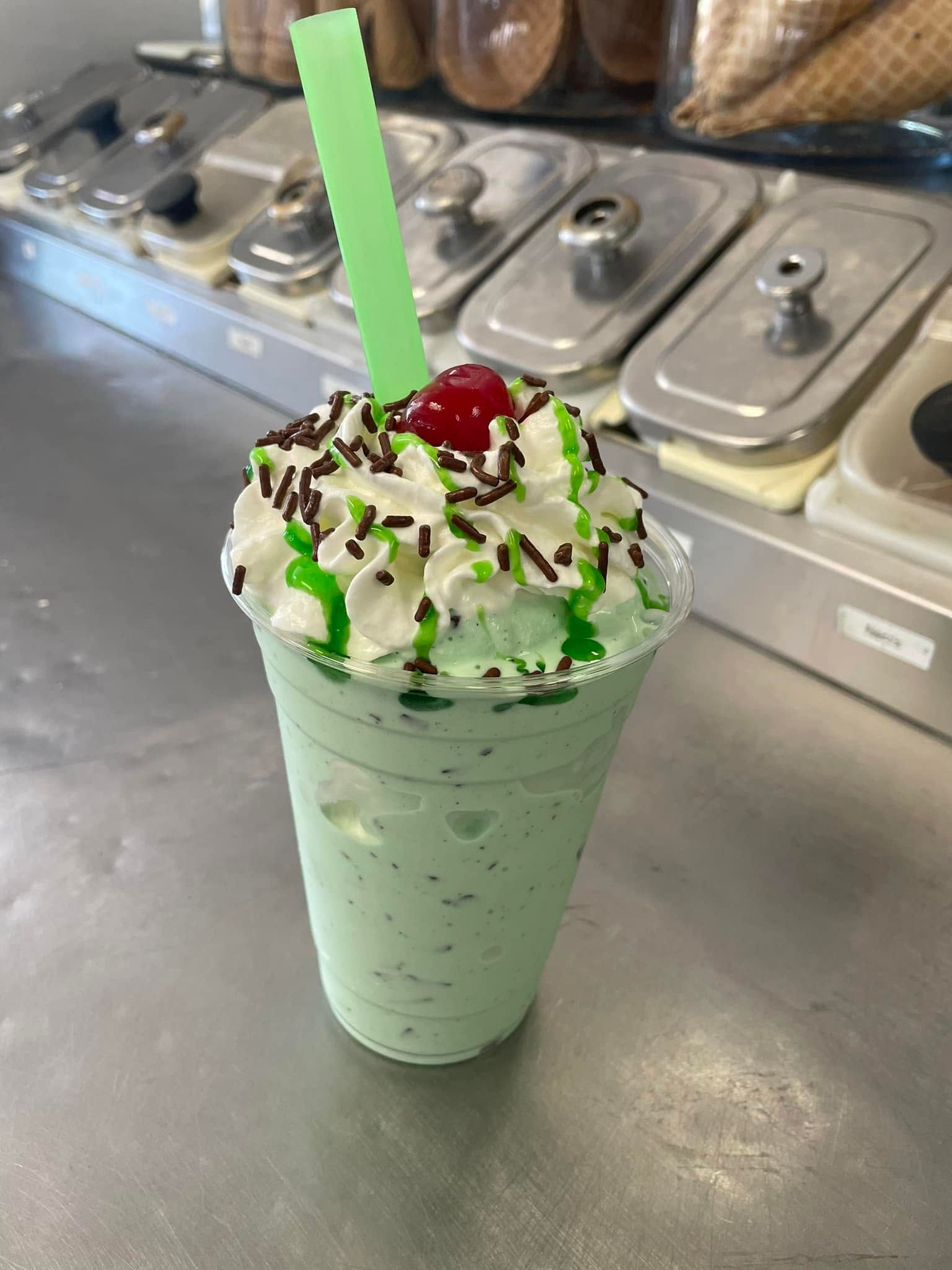 Mint milkshake topped with whipped cream, chocolate drizzle, and cherry on a counter with ice cream tubs