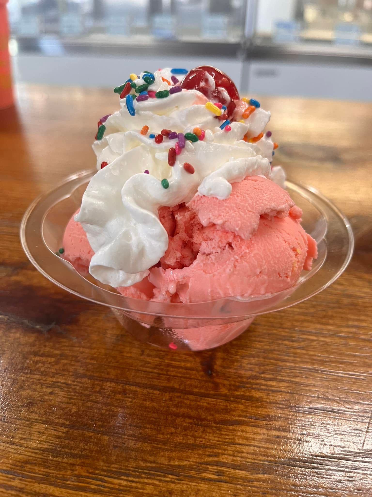 Pink ice cream sundae topped with whipped cream, sprinkles, and syrup in a clear bowl on a wooden table