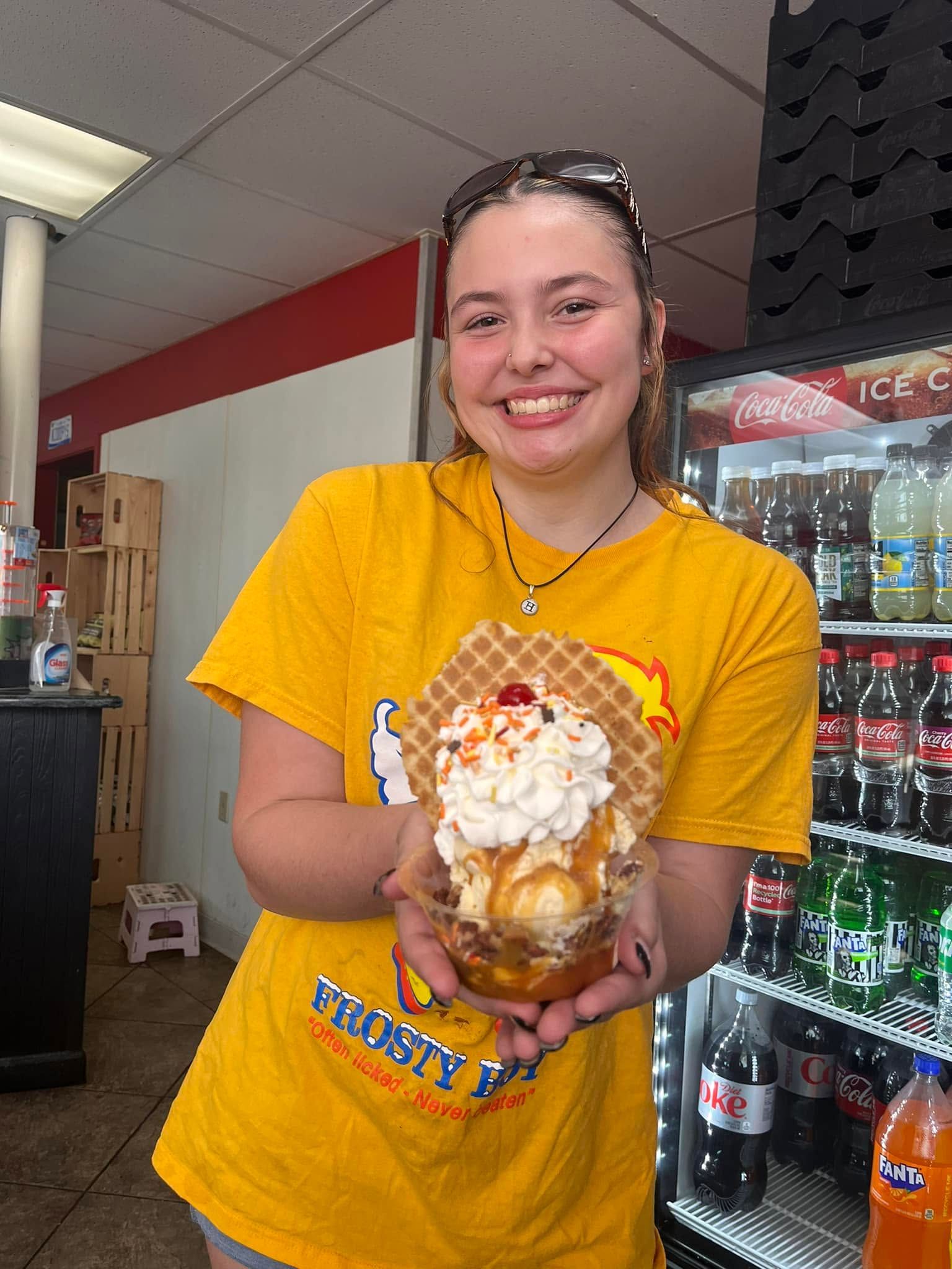 Smiling person in a yellow shirt holding a dessert topped with whipped cream in a convenience store.