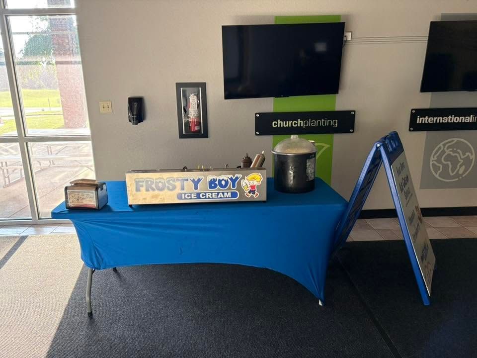 Church lobby table with blue tablecloth, Frosty Boy sign, candy jar, and wall-mounted TV.