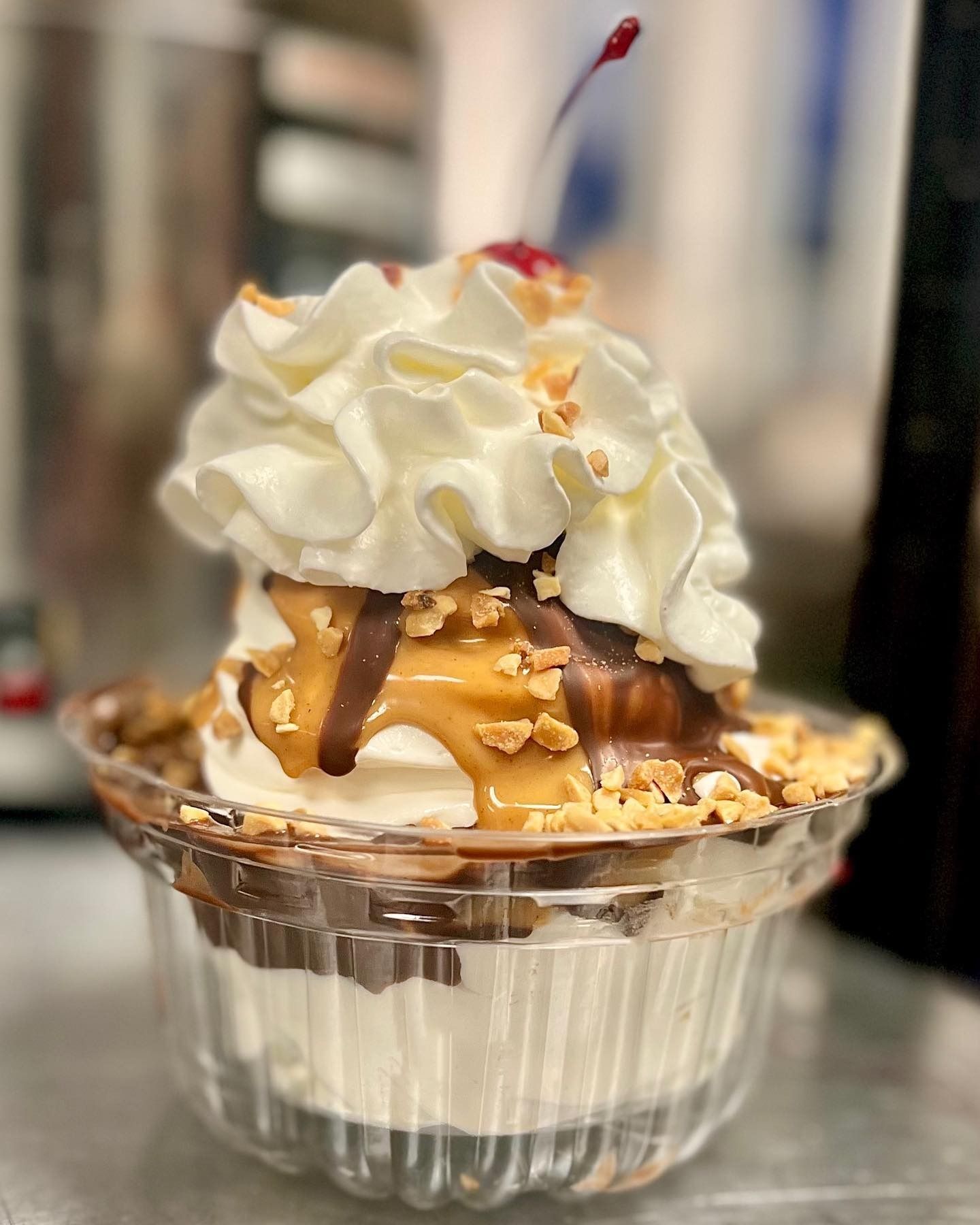 Ice cream sundae in a clear cup topped with whipped cream, caramel drizzle, nuts, and a cherry.