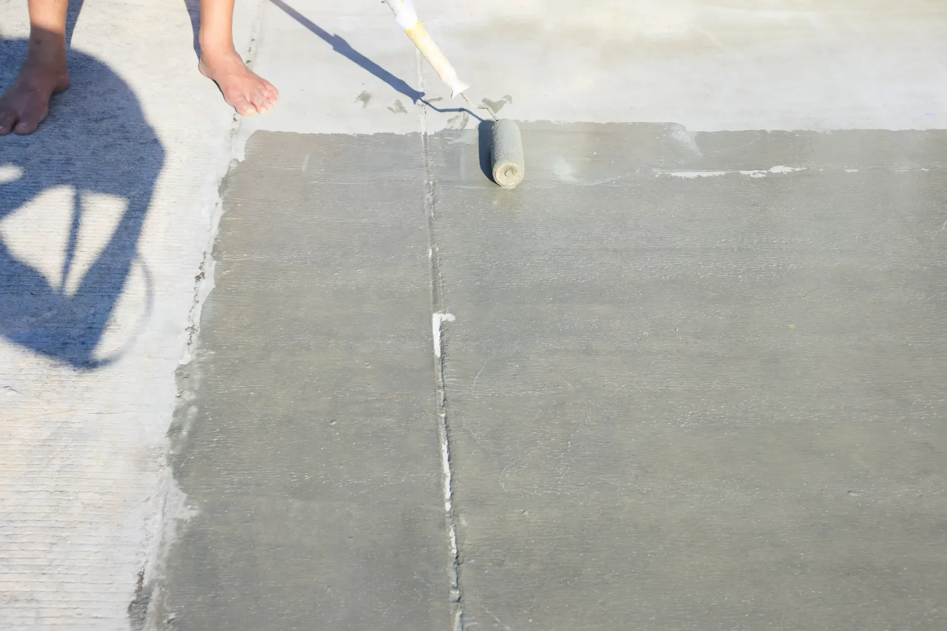 Person using a paint roller on concrete, applying a gray coating on an outdoor surface.