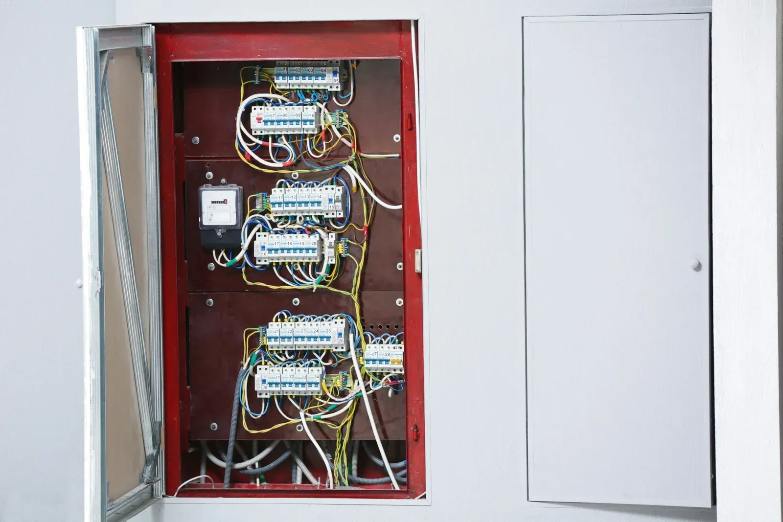 Open electrical panel on a white wall with wiring and circuit breakers, next to a closed white panel door.