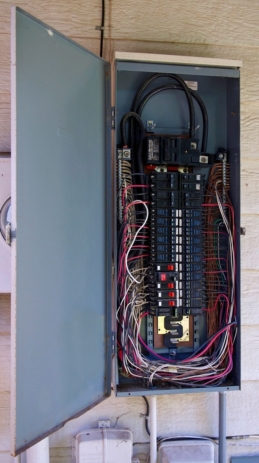 Open electrical breaker panel with exposed wiring and circuit breakers mounted on an exterior wall.