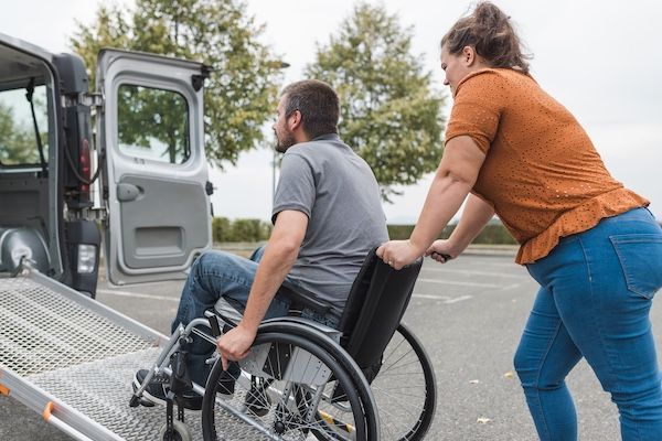A lady helping a man in a wheelchair into a medical transportation van