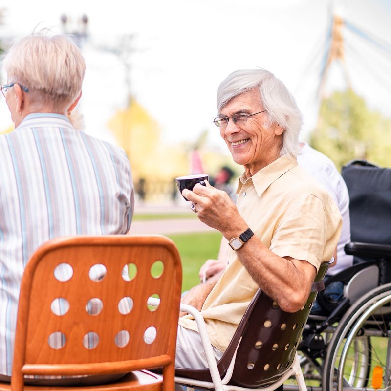 An elderly man in a wheelchair is drinking tea with a friend.