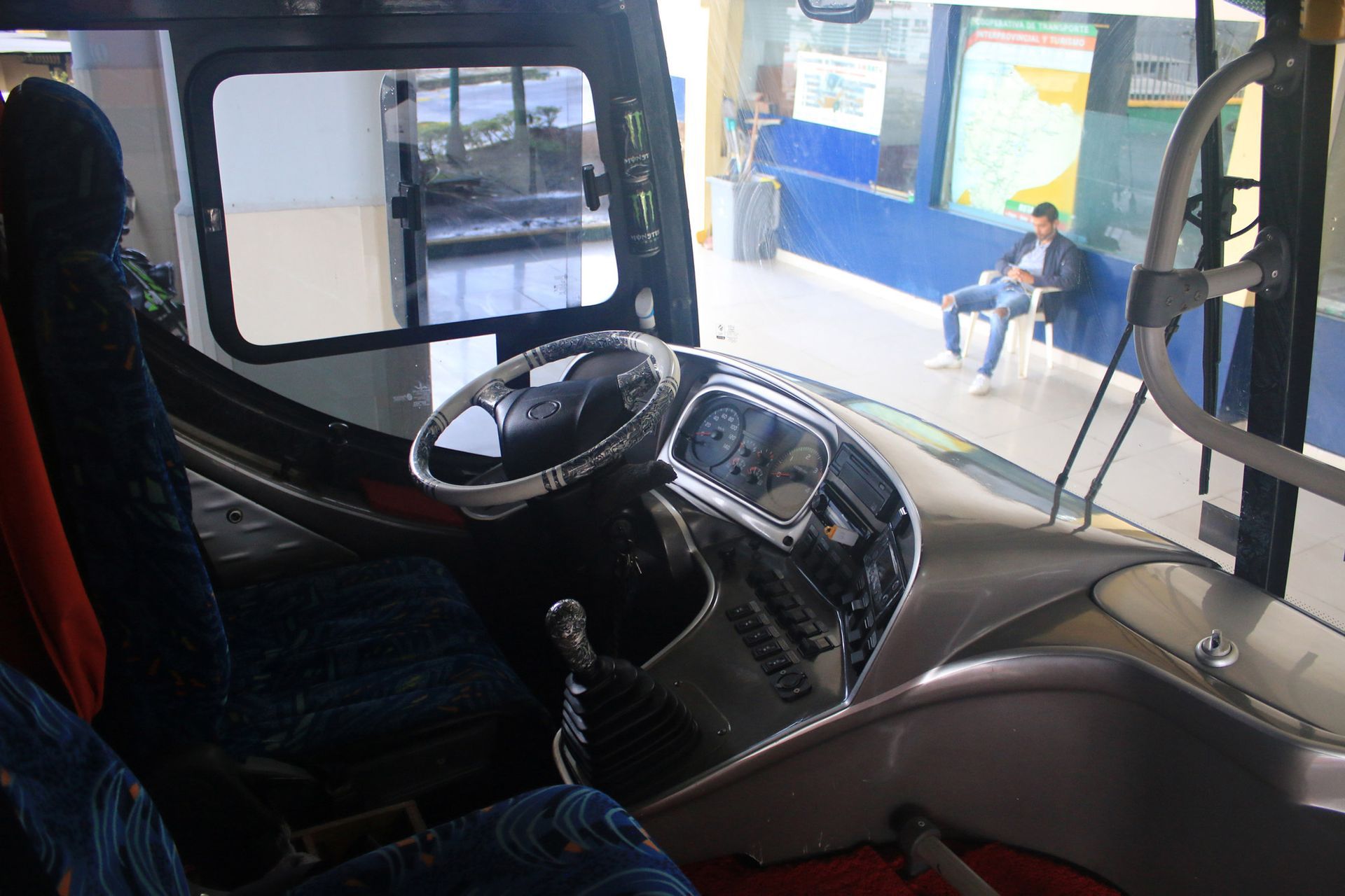 Interior bus cockpit with steering wheel and dashboard, looking towards a passenger waiting at a bus stop.