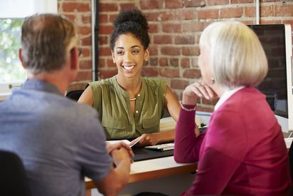 Woman smiling, speaking with a couple at a desk. Brick wall background.