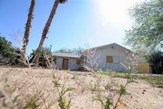 A weathered stucco house in a sunny, desert landscape. Dry brush in the foreground, palm trees.
