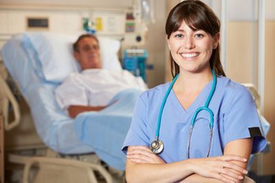 Nurse in blue scrubs, arms crossed, smiles in a hospital room with a patient in bed.