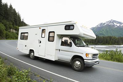 White RV driving on a road near a forest and mountains.