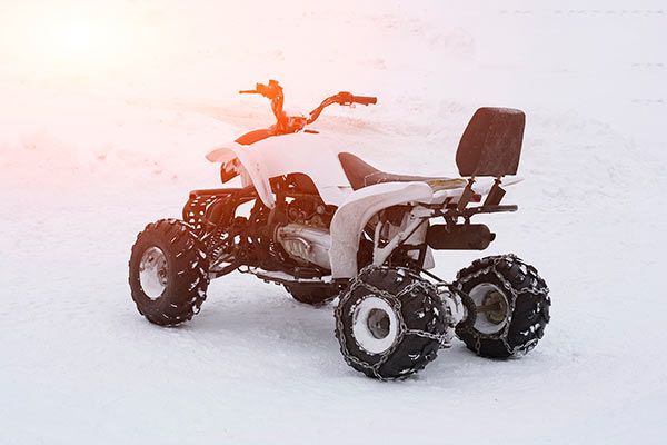 White ATV with snow chains on a snowy surface, bright sunlight overhead.