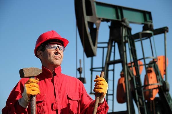 Oil worker in red overalls and hard hat holding tools, with oil pump in the background.