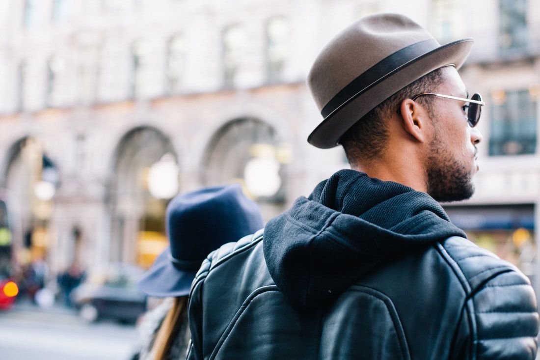 Man wearing a fedora and sunglasses in a city setting, with a leather jacket and another person in the background.