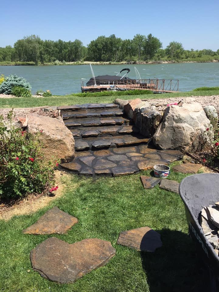 Stone steps lead through a garden to a lakeside dock with a boat, surrounded by flowers and grass.